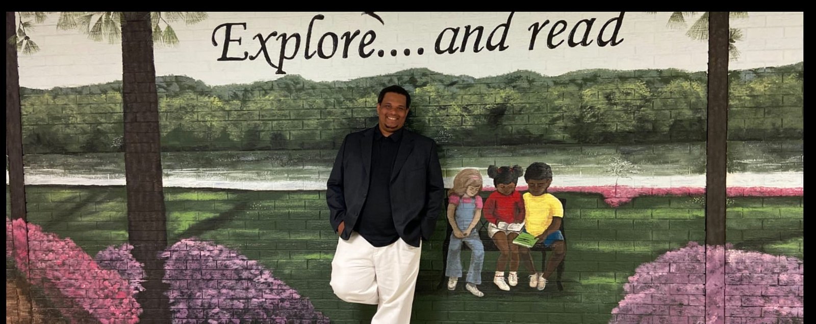 David D. Gibson Sr. standing in front of a painted mural of children reading on a bench, under the words “Explore.... and read”